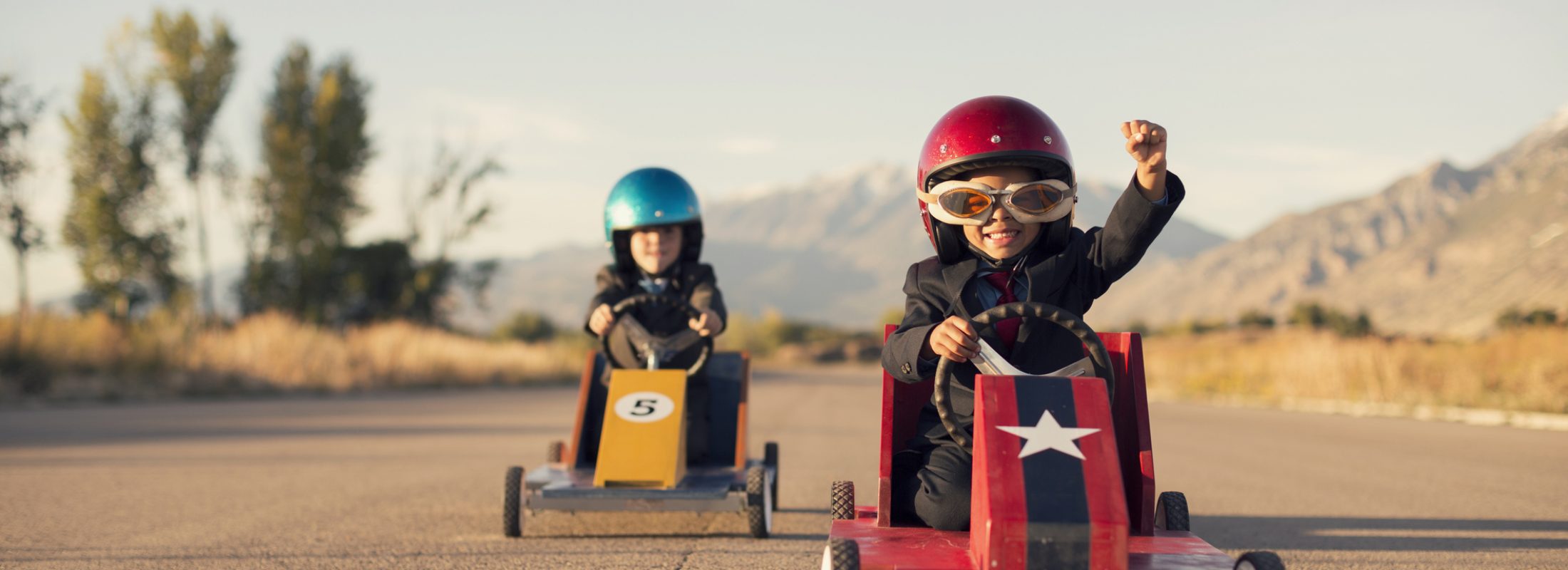 A young boy dressed as a businessman raises his arm in success as his homemade box car is in first place. Both boys are wearing helmets and goggles.